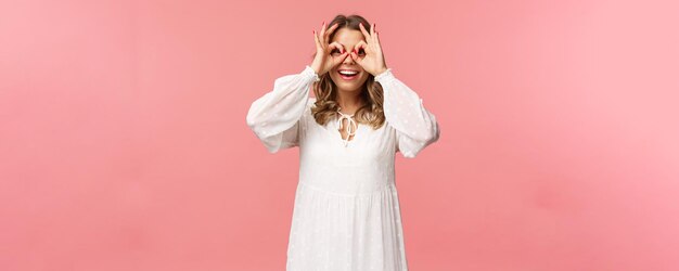 Portrait of dreamy cute and funny young blond girl seeing something interesting looking from okay signs as making glassmask with fingers over eyes smiling amused pink background