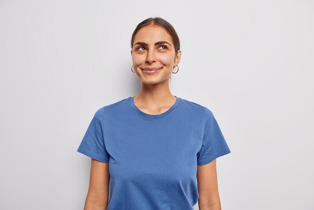 Portrait of dreamy dark haired woman smiles gently concentrated above thinks about something very pleasant recalls nice memories wears casual blue t shirt isolated over white studio wall