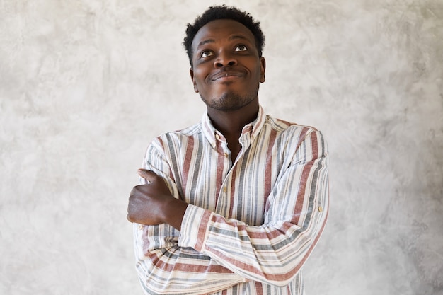 Портрет мечтательного задумчивого молодого афро-американского парня в полосатой рубашке, обнимающего себя и смотрящего вверх с задумчивым выражением лица, улыбающегося, мечтающего