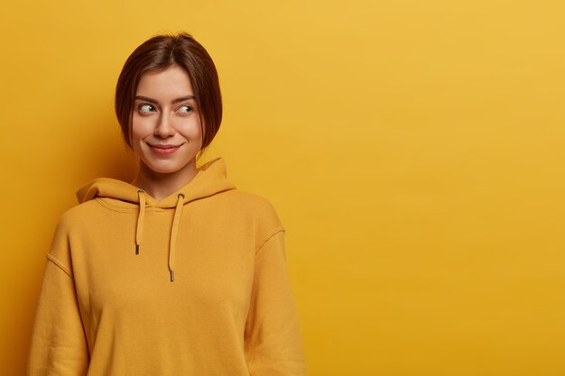 Portrait of dreamy satisfied young European woman looks aside on blank space, has pleasant look, wears hoodie, poses against yellow wall, empty space area, has intriguing plan in mind.