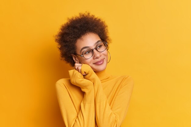 Portrait of dreamy twenty years old female keeps hands near face and has romantic expression has curly bushy hair wears spectacles turtleneck.