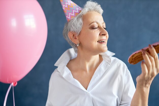 portrait of ecstatic happy retired lady in stylish white shirt and cone hat dancing to music at birthday party, holding pink helium balloon.