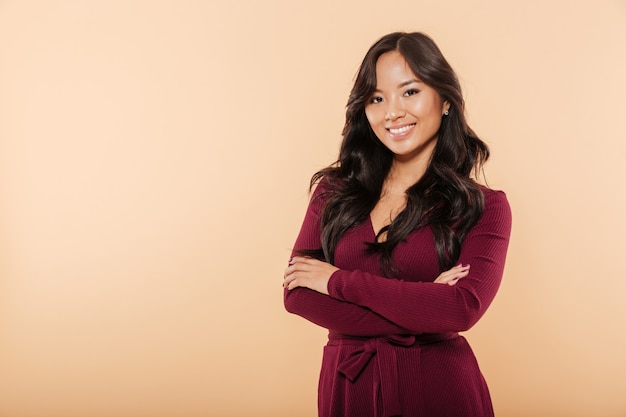 Portrait of elegant asian female in pretty maroon dress posing on camera with arms folded isolated over peach background