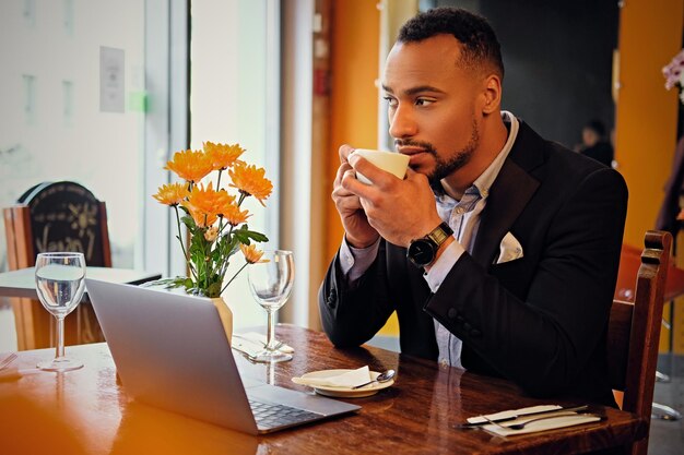 Portrait of elegant black American businessman dressed in a suit, using laptop and drinks coffee in a cafe.