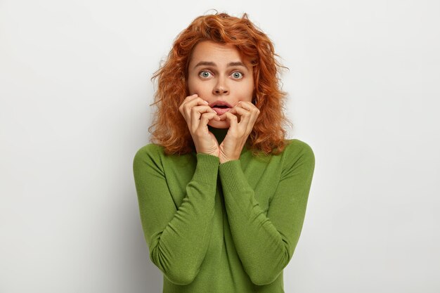 Portrait of embarrassed nervous woman has scared worried expression, keeps hands near opened mouth, afraids of hearing terrible news, has natural ginger hair, dressed in green casual clothes