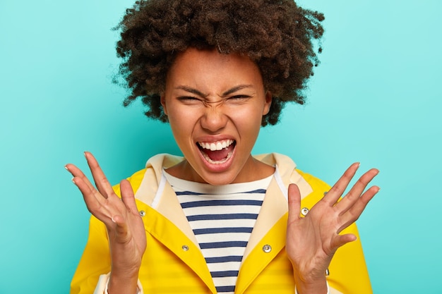 憂鬱な雨天に悩まされている感情的な怒っている巻き毛のアフロアメリカ人女性の肖像画、口を開いたまま、縞模様のジャンパーと黄色のレインコートを着て、青い背景に対してポーズをとる