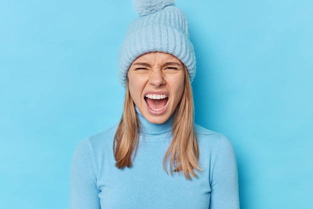 Portrait of emotional angry woman screams with widely opened mouth feels outraged expresses negative emotions wears turtleneck and hat isolated over blue background has quarrel with someone.