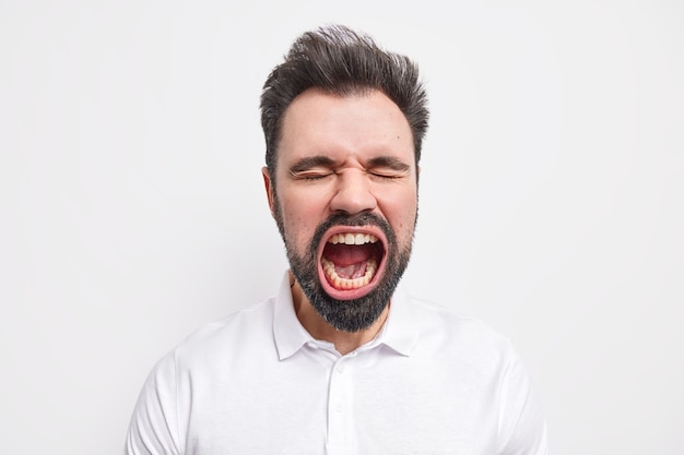 Portrait of emotional bearded mad European man keeps mouth wide opened closes eyes has thick beard dressed in shirt 