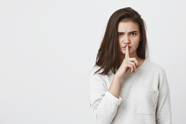 Portrait of emotional brunette woman dressed casually holding index finger on lips, saying 'shh', asking to keep silence or to keep her secret. Face expressions, emotions and feelings, body language