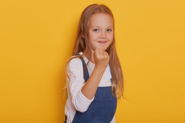 黄色の背景、彼女の拳を示すかわいい金髪の子供に分離された感情的な少女の肖像画