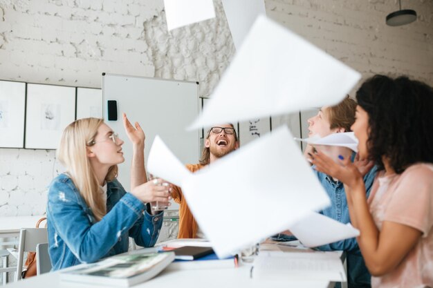 Portrait of emotional people happily throw up papers in office while working together Group of young students joyfully studying with books on table in classroom