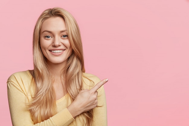 Portrait of emotional pleasant looking female has shining smile indicates with fore finger at blank copy space, advertizes something. Blonde attractive woman shows new product, attracts your attention