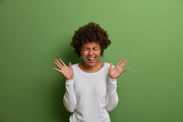 Портрет эмоционально позитивной темнокожей женщины громко смеется, веселится в помещении, поднимает ладони, держит глаза закрытыми, носит белый джемпер, выглядит беззаботно и расслабленно, изолированно на зеленой стене