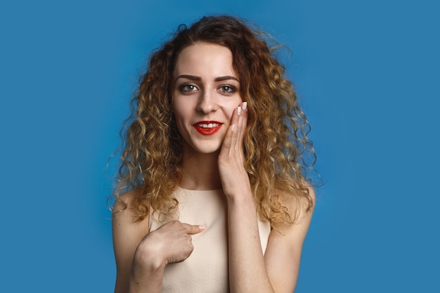 Portrait of emotional positive young Caucasian woman with loose curly hair posing , having excited look, smiling happily , touching face and pointing index finger at herself