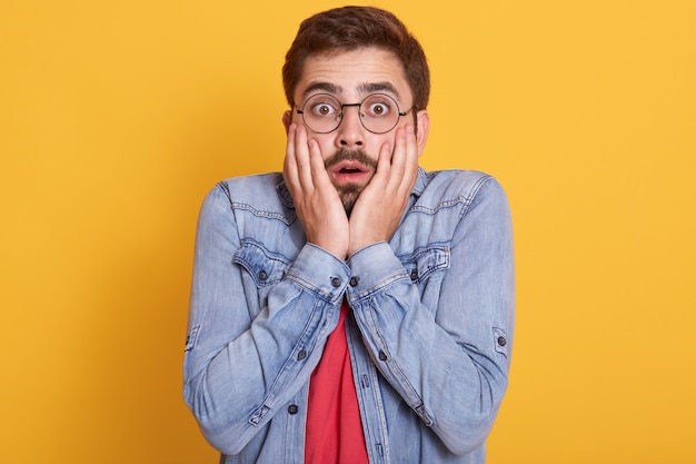 Portrait of emotional shocked scared guy opening eyes and mouth widely, putting hands to face, having impressed facial expression