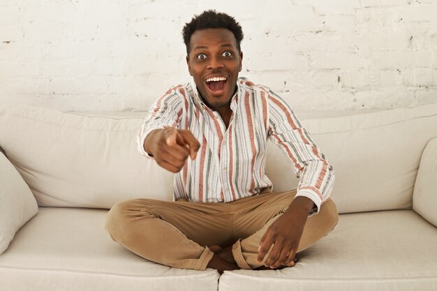 Portrait of emotional shocked young dark skinned guy relaxing at home, sitting comfortably on couch, having excited overjoyed look, pointing fore finger at camera