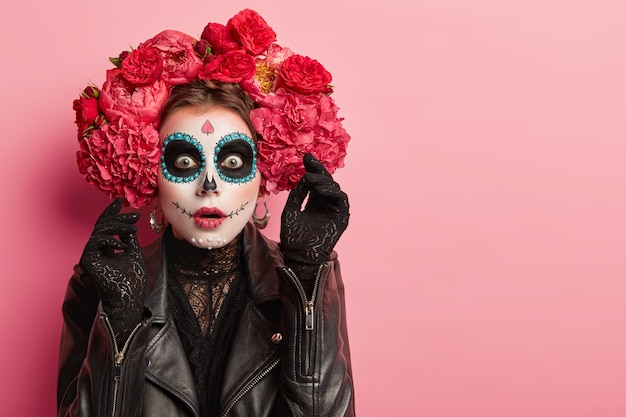 Portrait of emotional woman wears professional sugar skull makeup, has scared face expression, raises hand in black gloves with horror, prepares for Halloween holiday