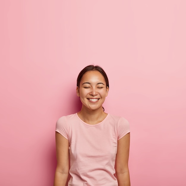 Портрет удовлетворенной эмоциями азиатской женщины с естественной красотой, темными причесанными волосами, счастливо улыбается, держит глаза закрытыми, носит повседневную футболку, изолированную на розовой стене. Люди, этническая принадлежность, положительные эмоции