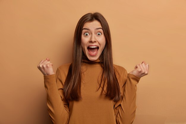 Portrait of emotive cheering woman with European appearance, clenches fists, finds out about her win, clenches fists with triumph, shares success