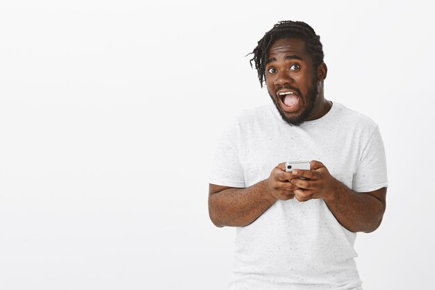 Portrait of emotive funny guy with braids posing against the white wall with his phone