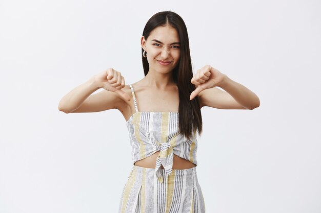 Portrait of emotive and playful cute female in matching top and shorts, showing thumbs down, wrinkling nose and smiling while flirting and expressing dislike or disappointment