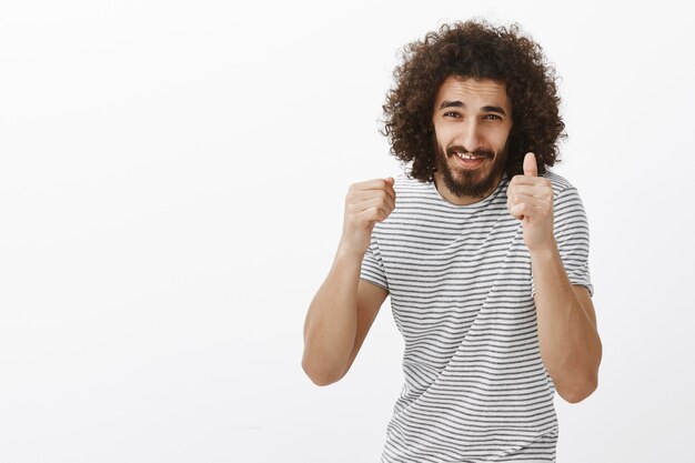 Portrait of energetic good-looking Eastern guy with curly haircut and beard, raising clenched fists and smiling with impatient expression