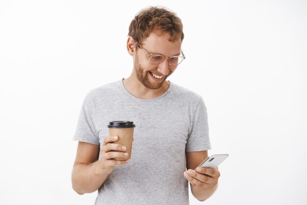 Portrait of entertained happy carefree guy with bristle holding paper cup and smartphone smiling broadly at gadget screen typing message