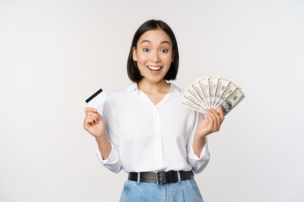 Портрет восторженной азиатской женщины, держащей деньги наличными и кредитной картой, улыбающейся, пораженной камерой на белом фоне