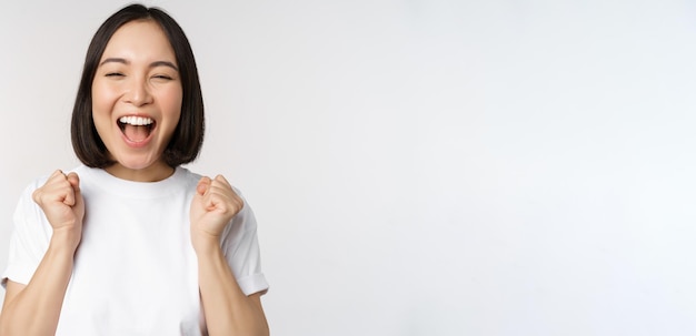 Portrait of enthusiastic asian woman winning celebrating and triumphing raising hands up achieve goa