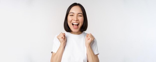 Portrait of enthusiastic asian woman winning celebrating and triumphing raising hands up achieve goal or success standing over white background