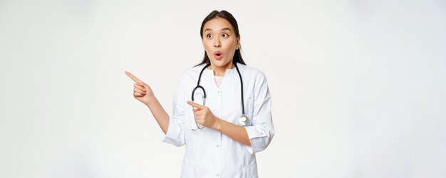 Portrait of enthusiastic female doctor asian physician pointing and looking left with surprised amaz