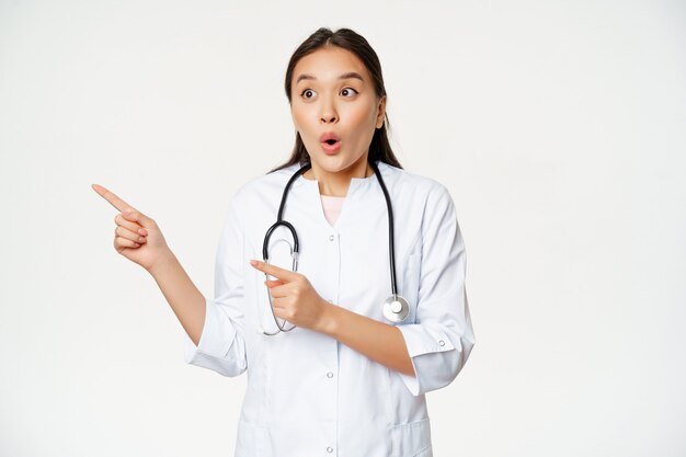 Портрет восторженной женщины-врача, азиатского врача, указывающего и смотрящего влево с удивленным, изумленным выражением лица, стоящего в медицинском халате на белом фоне.