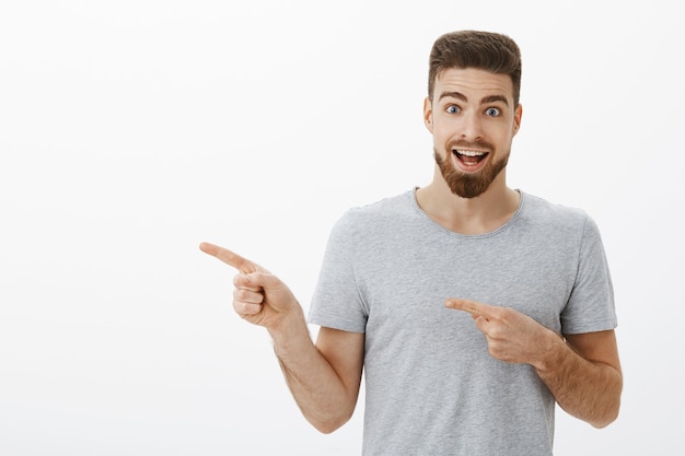 Free photo portrait of enthusiastic good-looking caucasian man pointing left and talking with amazement and excitement about perfect coworking space he found inviting to join over white wall