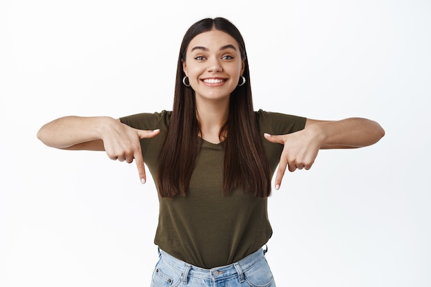 Ritratto di donna sorridente entusiasta che mostra pubblicità sul fondo, puntando le dita verso il basso e guardando felice davanti, in piedi sul muro bianco