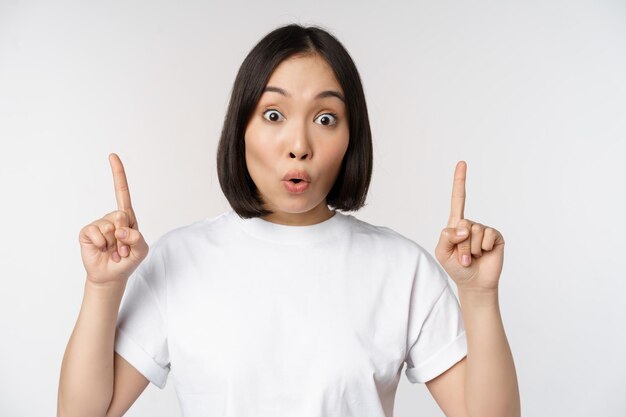 Portrait of enthusiastic young woman asian girl smiling pointing fingers up showing advertisement upwards standing over white background