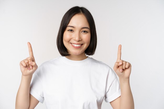 Portrait of enthusiastic young woman asian girl smiling pointing fingers up showing advertisement upwards standing over white background