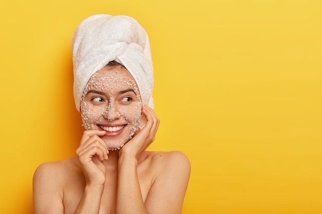 Portrait of European woman applies organic facial mask for cleaning skin, cares about complexion, smiles gently, shows white teeth, has bare shoulders, stands against yellow wall with blank space