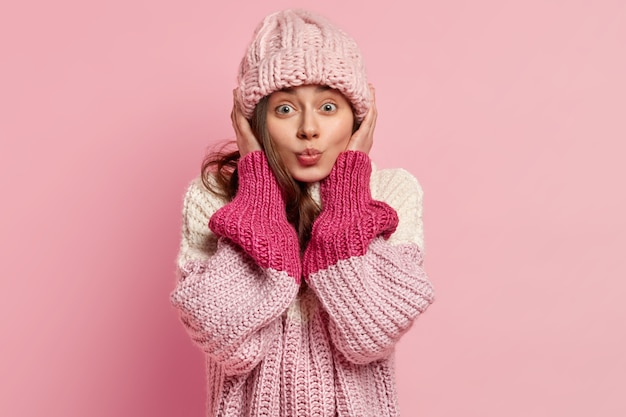 Portrait of European woman wears knitted headgear, dressed in loose sweater, wants to kiss someone, isolated over pink wall. Casual style. Winter fashion concept. Human facial expressions