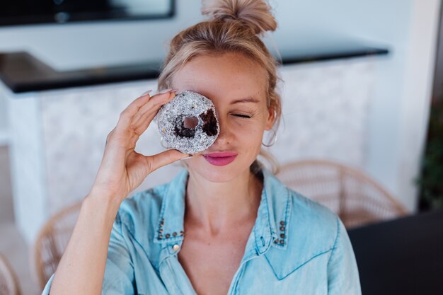 Portrait of european woman with blonde hair enjoying donuts in kitchen at home villa.