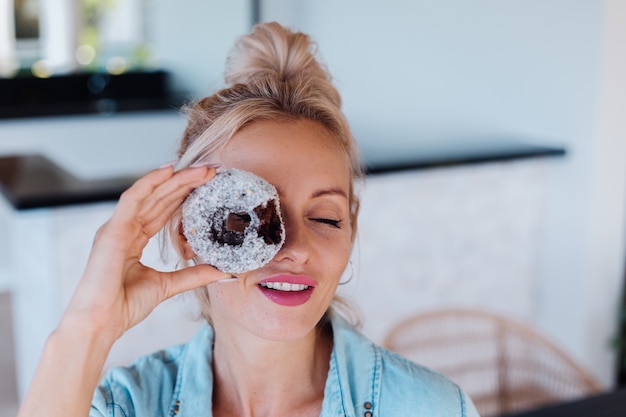 Портрет европейской женщины со светлыми волосами, наслаждаясь пончиками на кухне на домашней вилле.