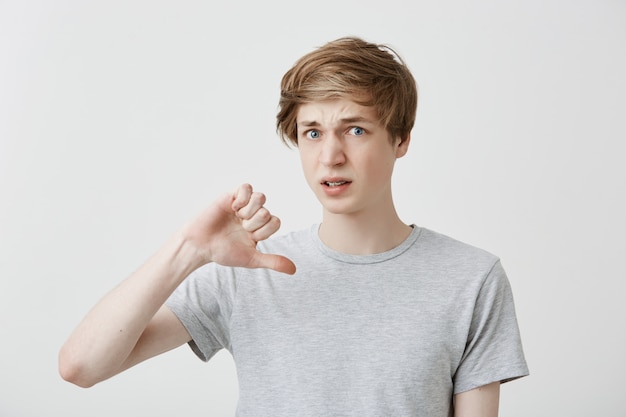 Portrait of european young man keeps thumb down, disapproves something, frowns face in discontent. Good looking fair-haired guy wears gray t-shirt dislikes film and doesn`t recommend to watch it