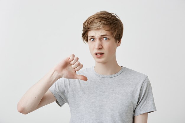 Портрет европейского молодого человека держит большой палец вниз, что-то не одобряет, недовольно хмурится. Красивый светловолосый парень в серой футболке не любит фильм и не рекомендует смотреть его