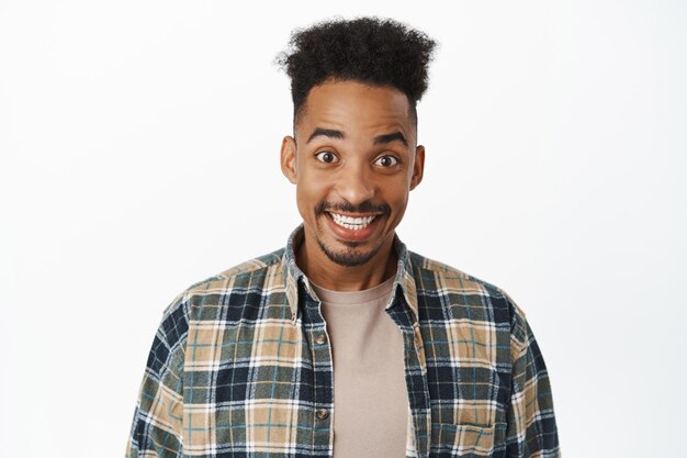 Portrait of excited african american man smiling white teeth, looking thrilled and amused, staring at something cool, standing in checked shirt against white background.