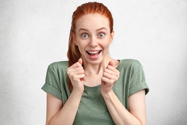 Portrait of excited amazed beautiful young female with attractive look, has overjoyed expression, gestures actively and clenches fists, dressed casually, isolated over white