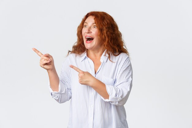 Portrait of excited and amazed, happy middle-aged woman react to thrilling promo banner, pointing and looking upper left corner fascinated, standing upbeat over white background.