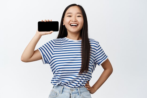 Портрет возбужденной азиатской девушки показывает горизонтальный экран своего мобильного телефона, довольно улыбается и рекомендует приложение на белом