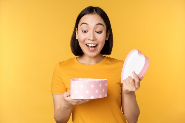 Portrait of excited asian woman open gift box with surprised happy face standing over yellow background