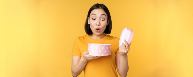 Portrait of excited asian woman open gift box with surprised happy face standing over yellow background