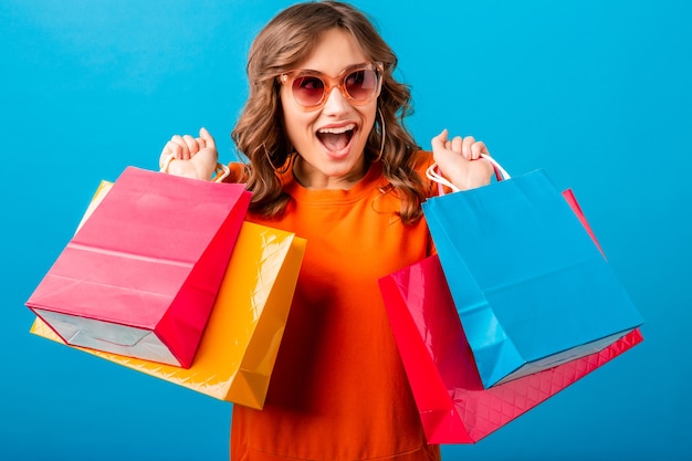 Portrait of excited attractive smiling stylish woman shopaholic in orange trendy dress holding shopping bags on blue studio background isolated