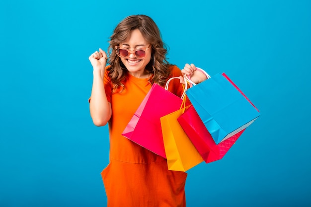 Portrait of excited attractive smiling stylish woman shopaholic in orange trendy dress holding shopping bags on blue studio background isolated